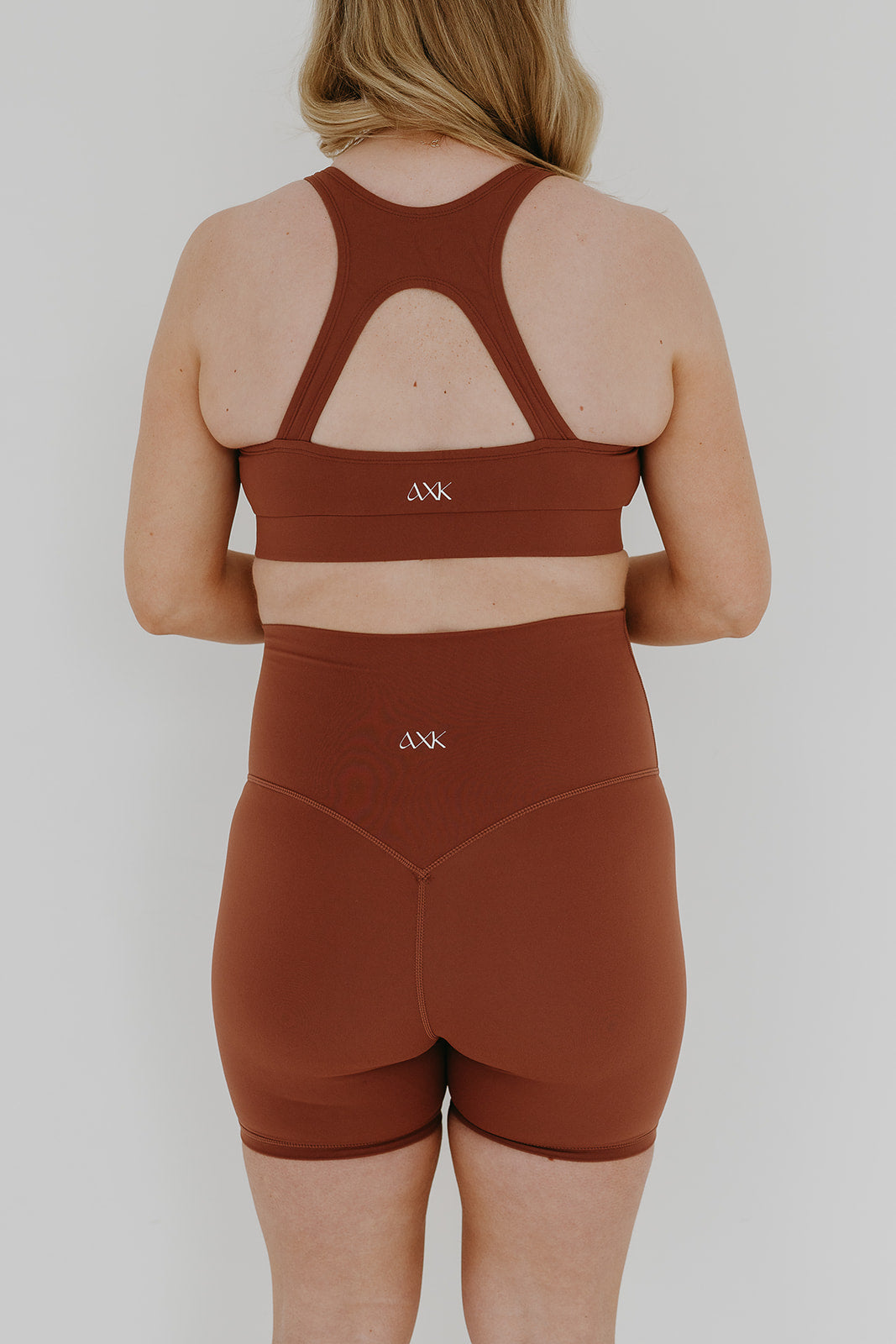 Back view of pregnant woman wearing our best reviewed premium brown maternity bike shorts with a supportive high waist and matching high-impact, racerback maternity and nursing sports bra, standing against a white background. Premium maternity athletic and activewear by AXK - high quality petite, tall, and plus size maternity.