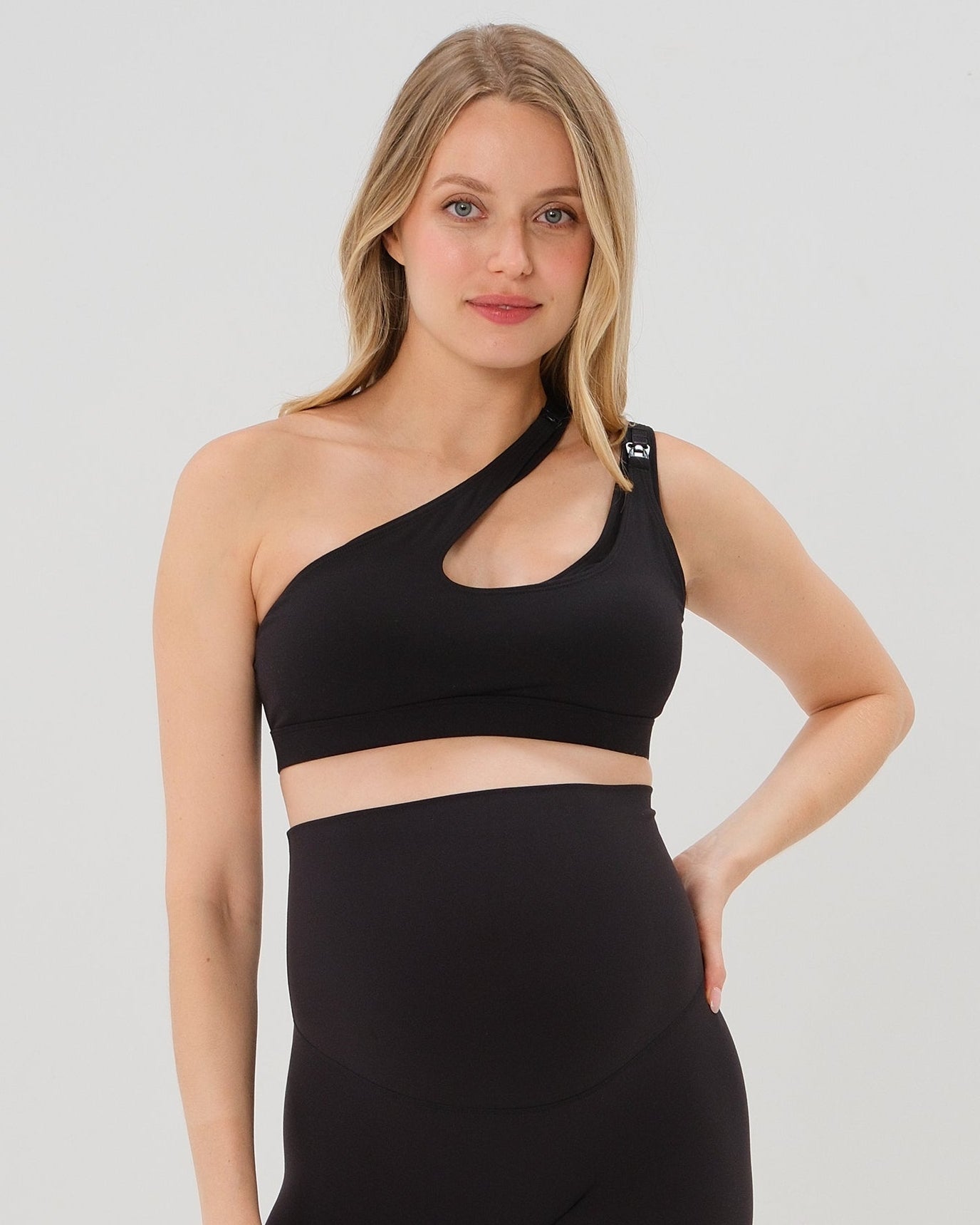 Pregnant woman in a one-shoulder black maternity and nursing sports bra, wearing matching black maternity leggings. Best selling 2026 premium maternity athletic and activewear by AXK, offered in regular and plus sizes, with glowing 5 star reviews.
