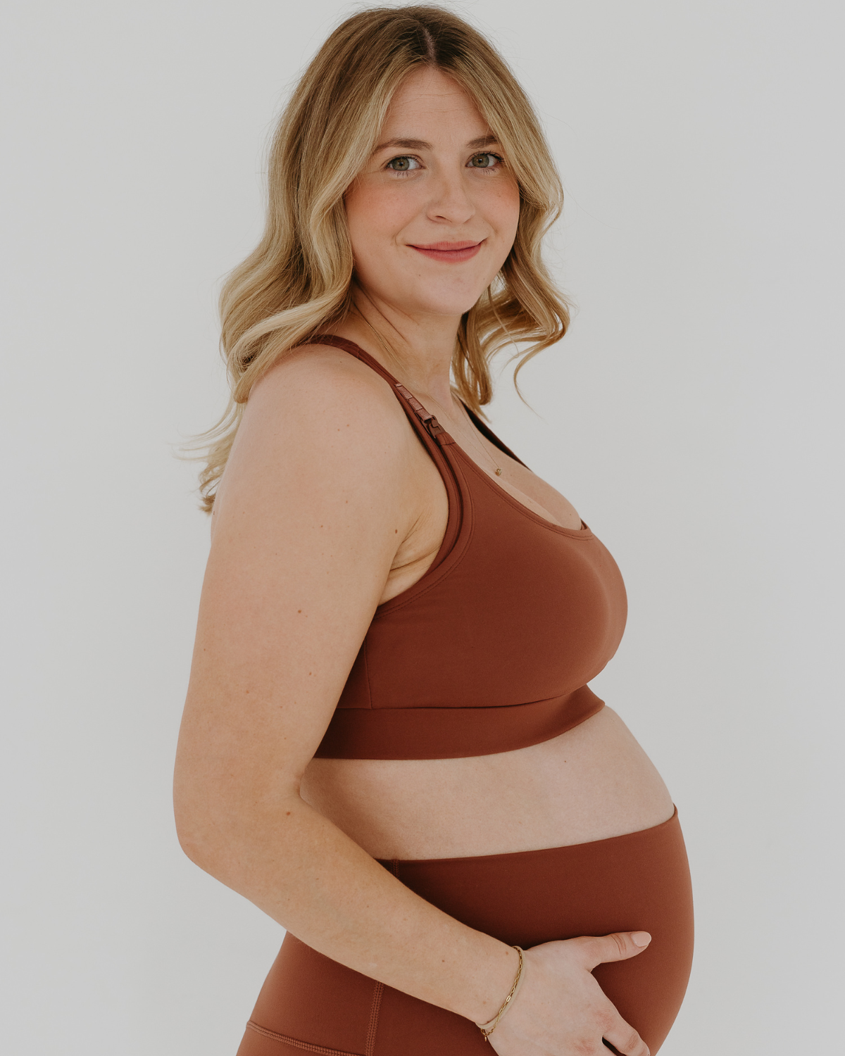 Side view of a smiling pregnant woman wearing a brown high-impact, racerback, maternity and nursing sports bra and matching high-waist maternity leggings. She has her hands cradling her belly standing in front of a white background. Premium maternity athletic and activewear by AXK.