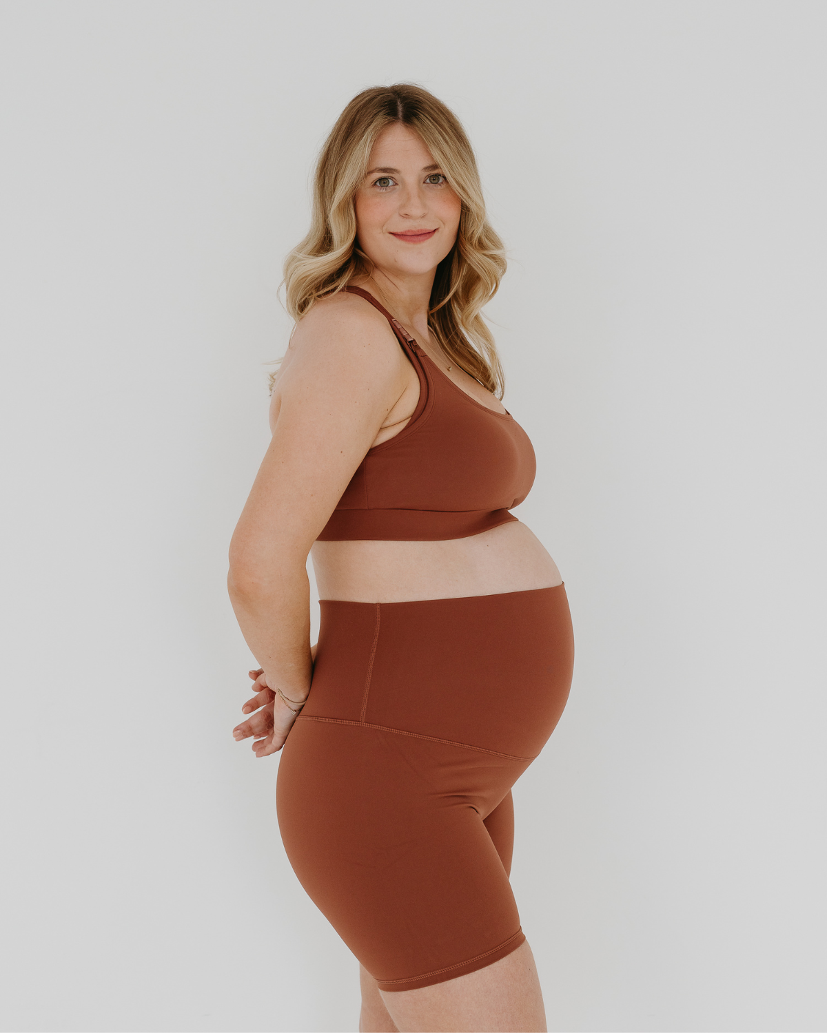 Pregnant woman wearing premium brown maternity bike shorts with a supportive high waist and matching high-impact, racerback maternity and nursing sports bra, standing against a white background. Premium maternity athletic and activewear by AXK.