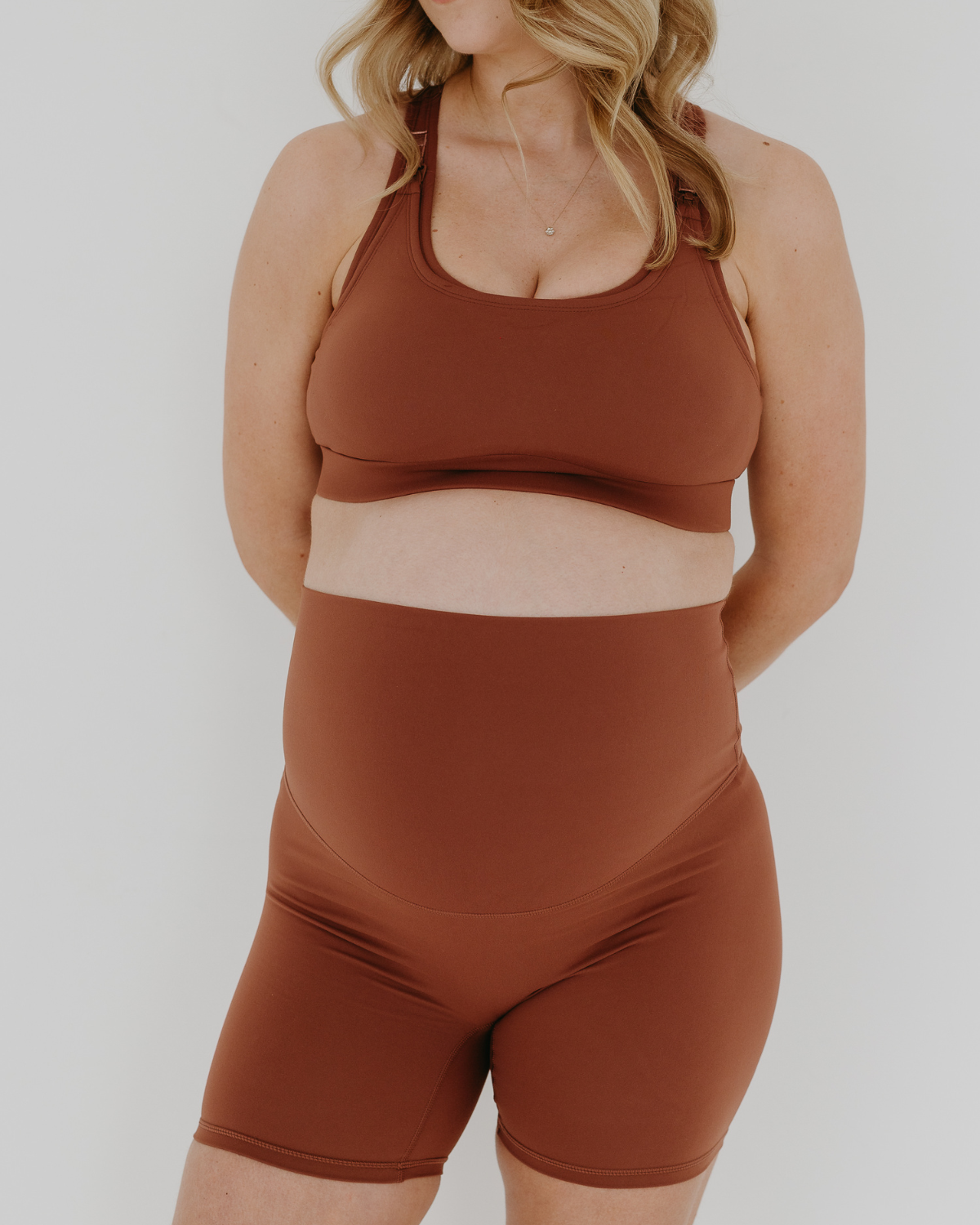 Pregnant woman wearing our best reviewed premium brown maternity bike shorts with a supportive high waist and matching high-impact, racerback maternity and nursing sports bra, standing against a white background. Premium maternity athletic and activewear by AXK - high quality petite, tall, and plus size maternity.