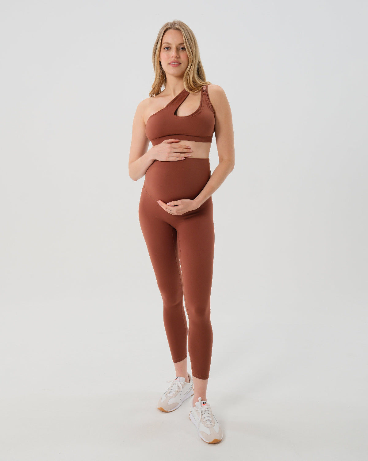 Pregnant woman wearing our best reviewed premium brown maternity leggings for 2026 with a flattering scrunch butt and matching trendy one shoulder nursing sports bra, standing against a white background. Premium maternity athletic and activewear by AXK.