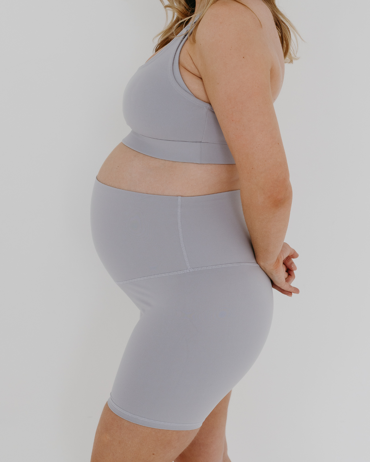 Pregnant woman wearing premium light grey maternity bike shorts with a supportive high waist and matching high-impact, racerback maternity and nursing sports bra, standing against a white background. Premium maternity athletic and activewear by AXK.