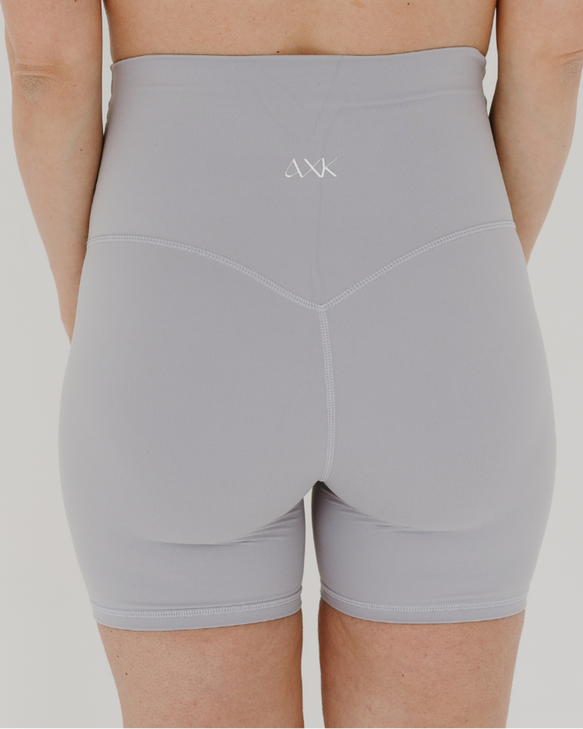Back view of a pregnant woman wearing premium light grey maternity bike shorts with a supportive high waist and matching high-impact, racerback maternity and nursing sports bra, standing against a white background. Premium maternity athletic and activewear by AXK.