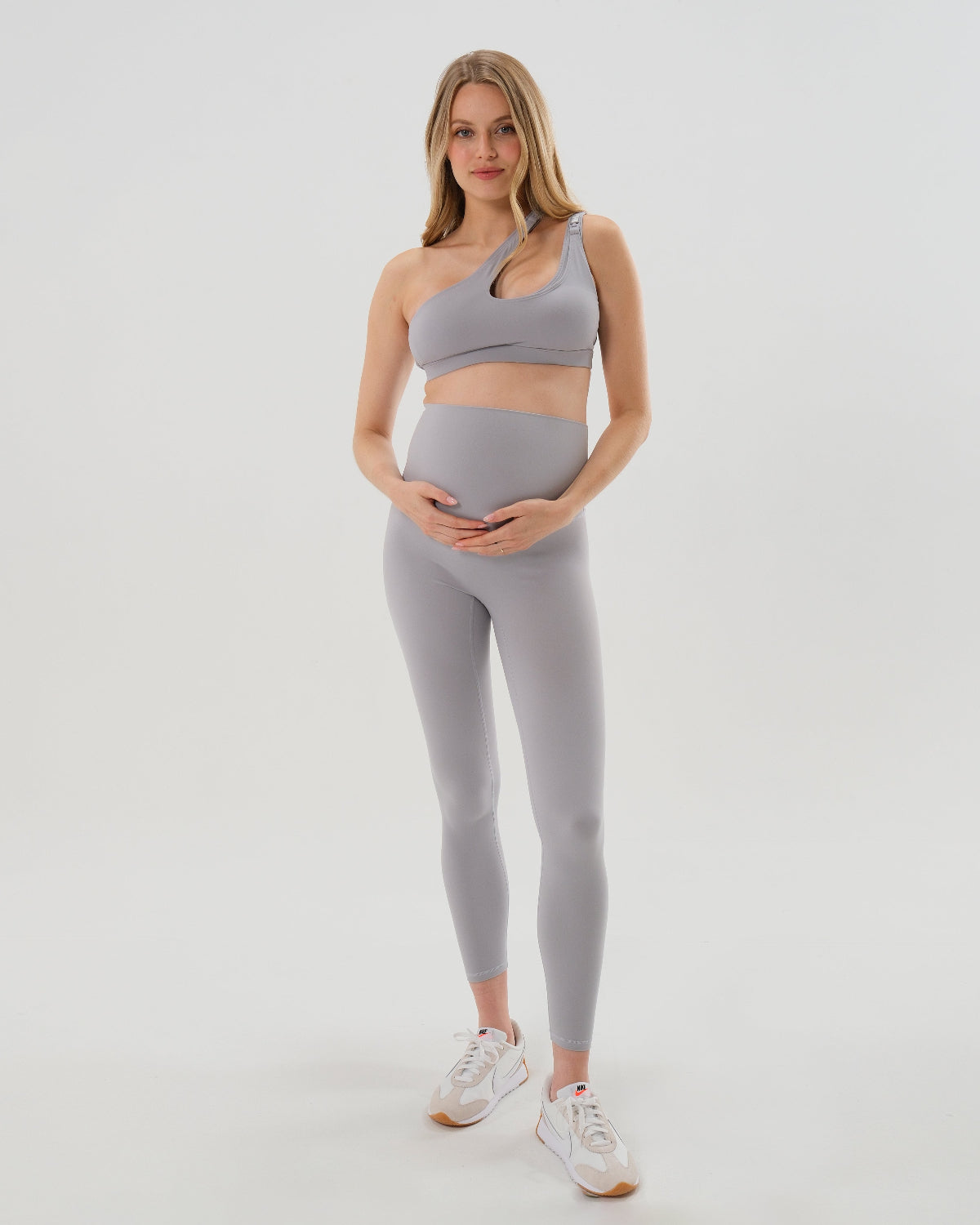 Pregnant woman wearing our best reviewed premium brown maternity leggings for 2026 with a flattering scrunch butt and matching trendy one shoulder nursing sports bra, standing against a white background. Premium maternity athletic and activewear by AXK.