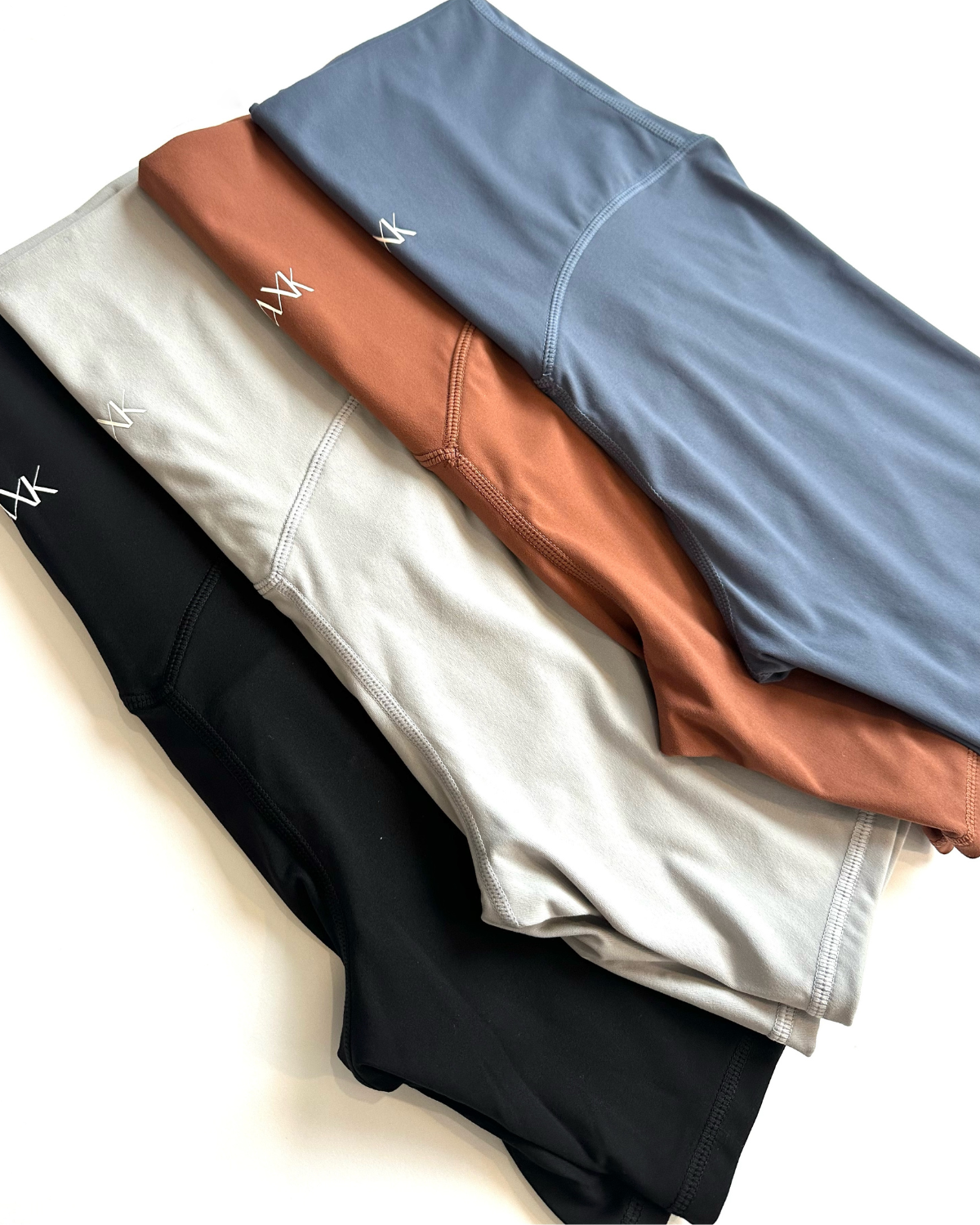 Stack of folded premium maternity bike shorts in black, light grey, brown, and blue, featuring AXK logo detail, displayed on a white background. Premium maternity athletic and activewear by AXK.