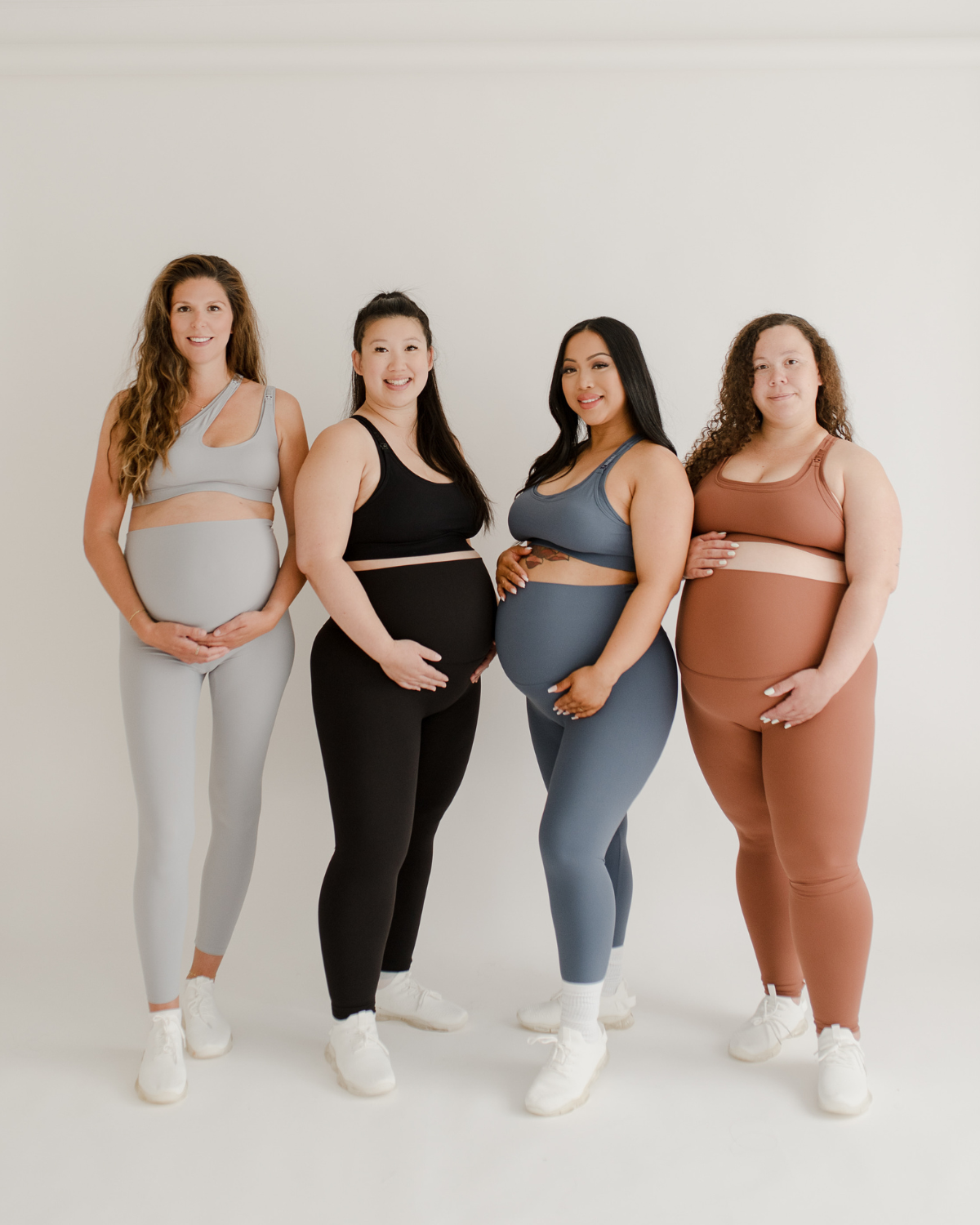 Four pregnant women wearing premium maternity leggings with flattering scrunch butt and matching high-impact, racerback nursing sports bras in light grey, black, blue, and brown, standing together against a white background. Premium maternity athletic and activewear by AXK.