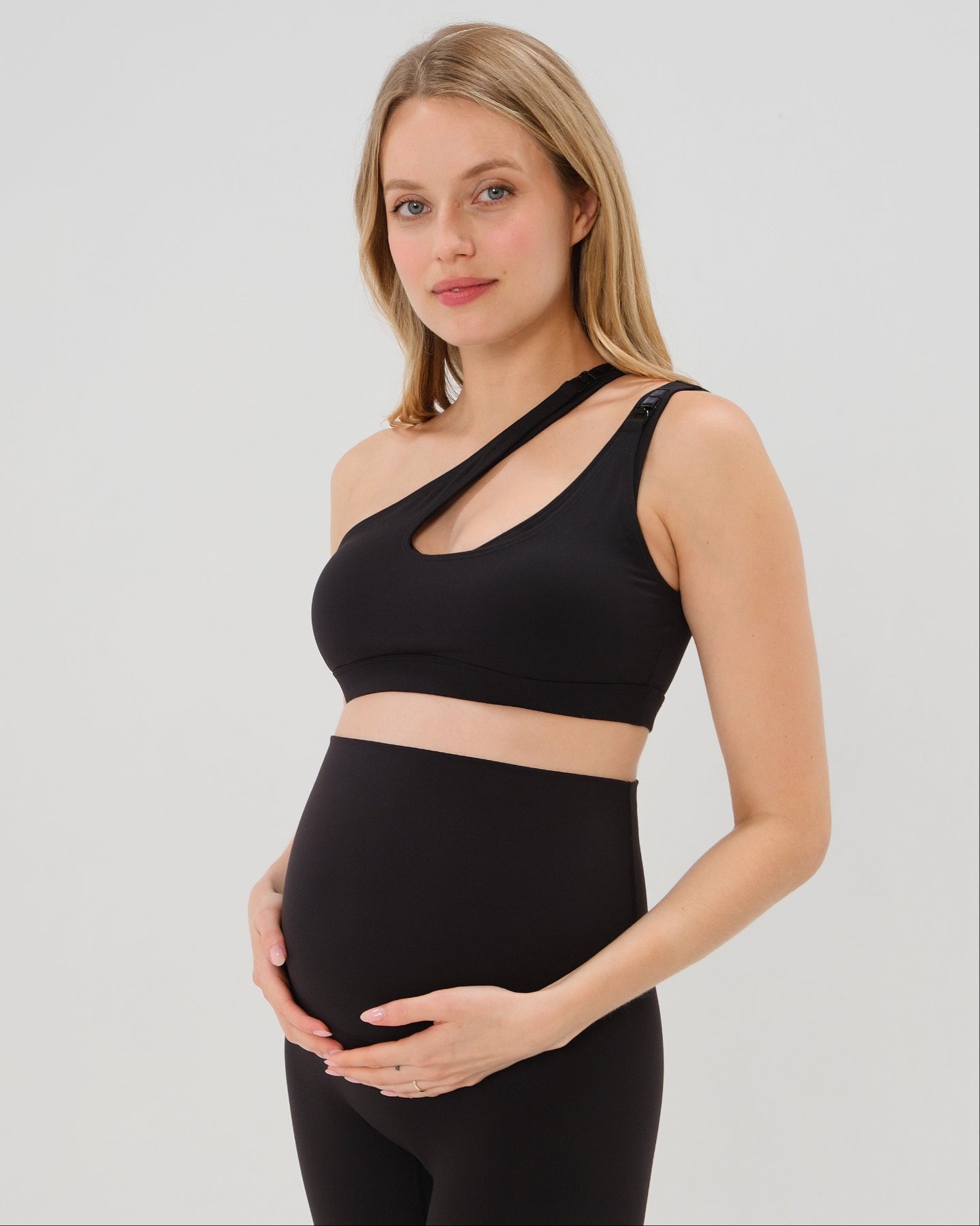 Pregnant woman in a one-shoulder black maternity and nursing sports bra, wearing matching black maternity leggings. Best selling 2026 premium maternity athletic and activewear by AXK, offered in regular and plus sizes, with glowing 5 star reviews. 