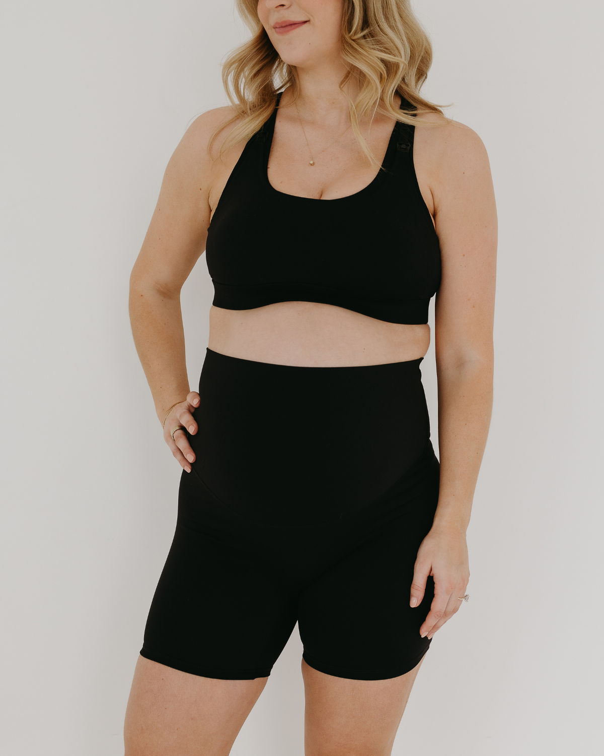 Pregnant woman wearing premium black maternity bike shorts with a supportive high waist and matching high-impact, racerback maternity and nursing sports bra, standing against a white background. Premium maternity athletic and activewear by AXK.