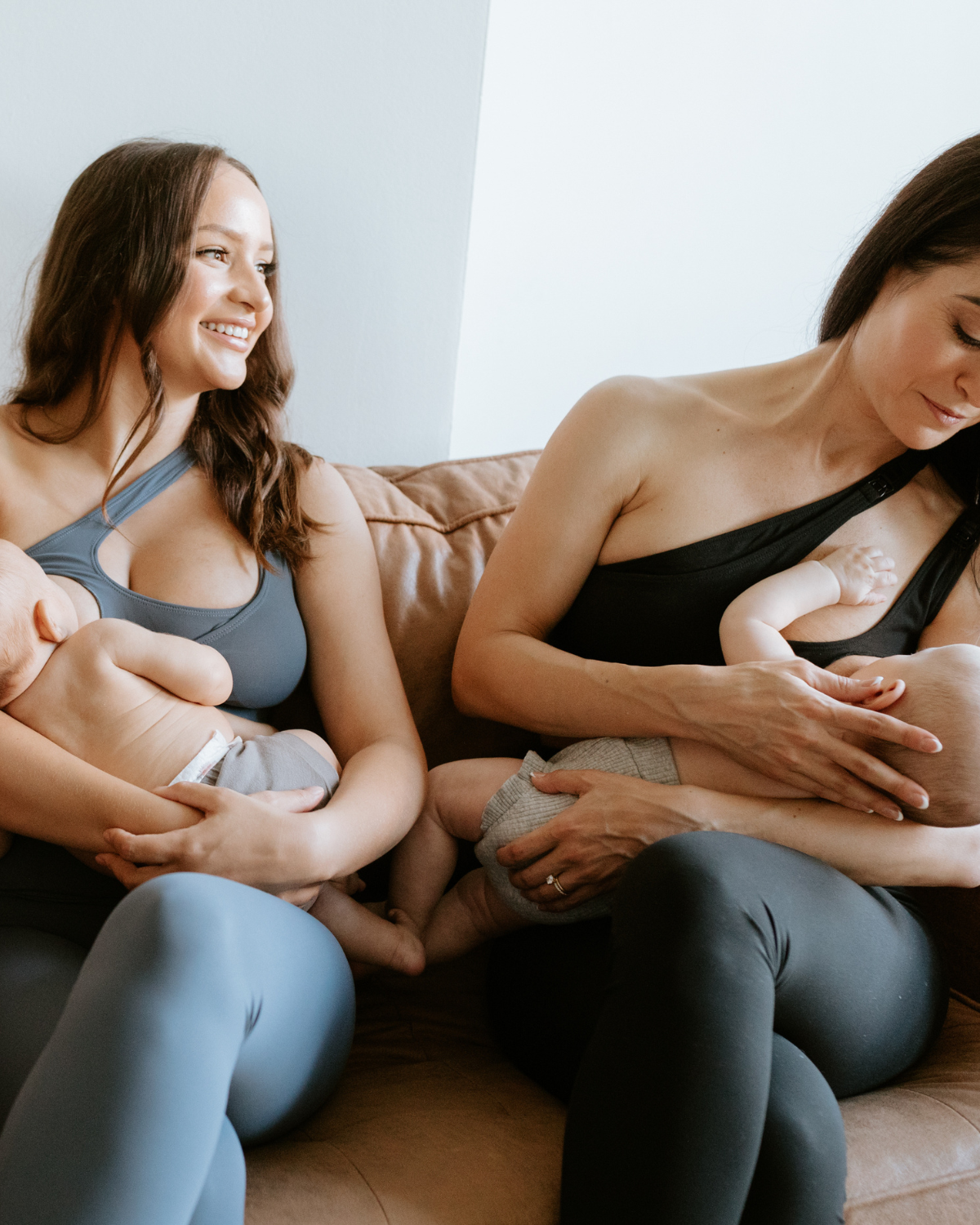 Two mothers sitting on a couch, each breastfeeding their baby while wearing one-shoulder, low-impact maternity and nursing sports bras by AXK. One bra is black and the other is blue, both designed with pull-down nursing access for style and function. Premium maternity activewear by AXK