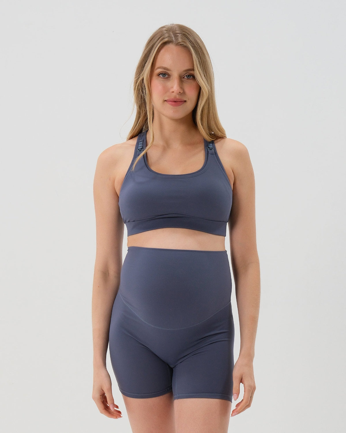 Pregnant woman wearing our premium blue best reviewed maternity bike shorts  for 2026 with a supportive high waist and matching high-impact, racerback maternity and nursing sports bra, standing against a white background. Premium maternity athletic and activewear by AXK.