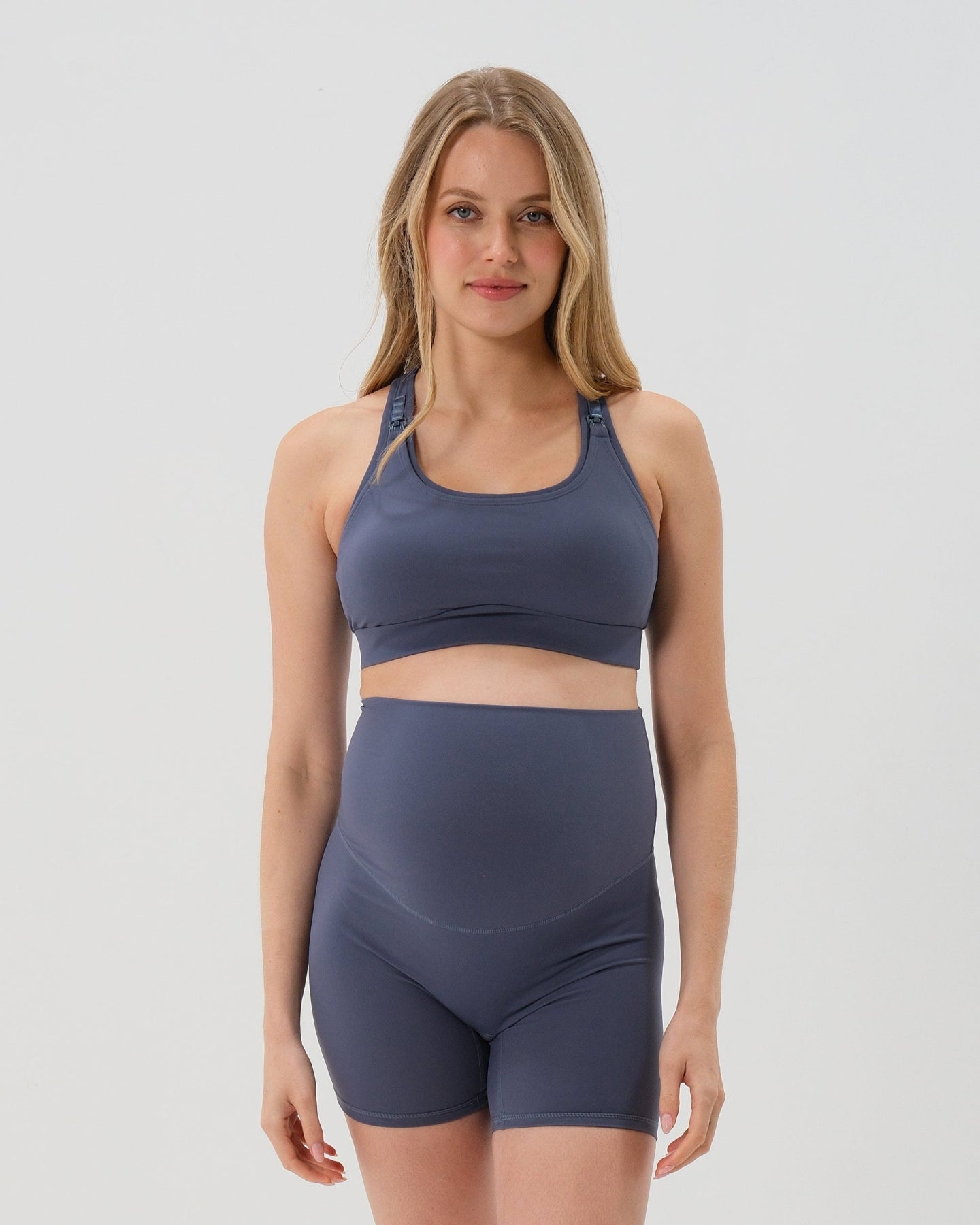 Pregnant woman wearing premium blue maternity bike shorts with a supportive high waist and matching high-impact, racerback maternity and nursing sports bra, standing against a white background. Premium maternity athletic and activewear by AXK - high quality petite, tall, and plus size maternity. 