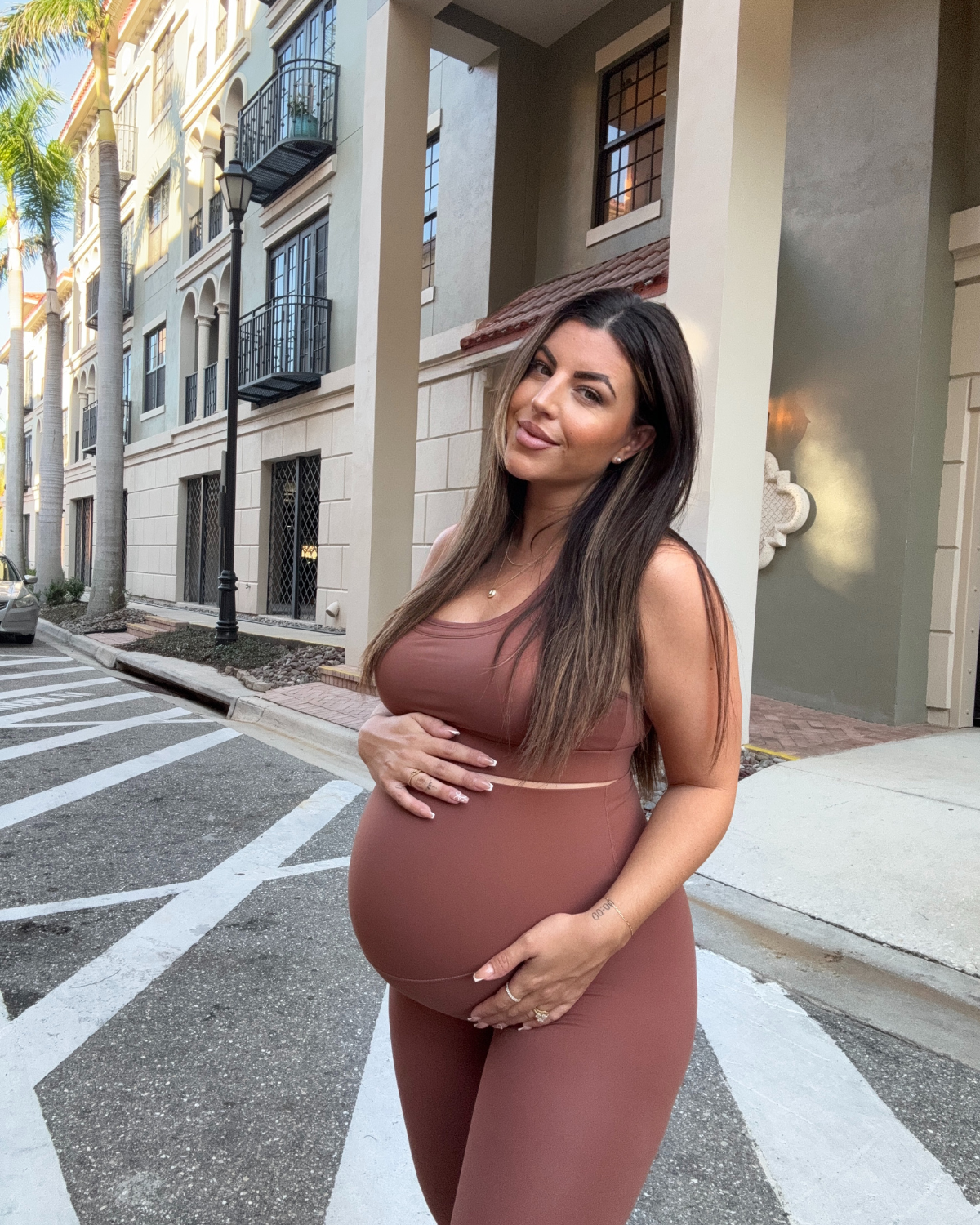 Pregnant woman wearing premium brown maternity leggings with a flattering scrunch butt and matching high-impact, racerback nursing sports bra, standing outdoors in an urban setting. Premium maternity athletic and activewear by AXK.