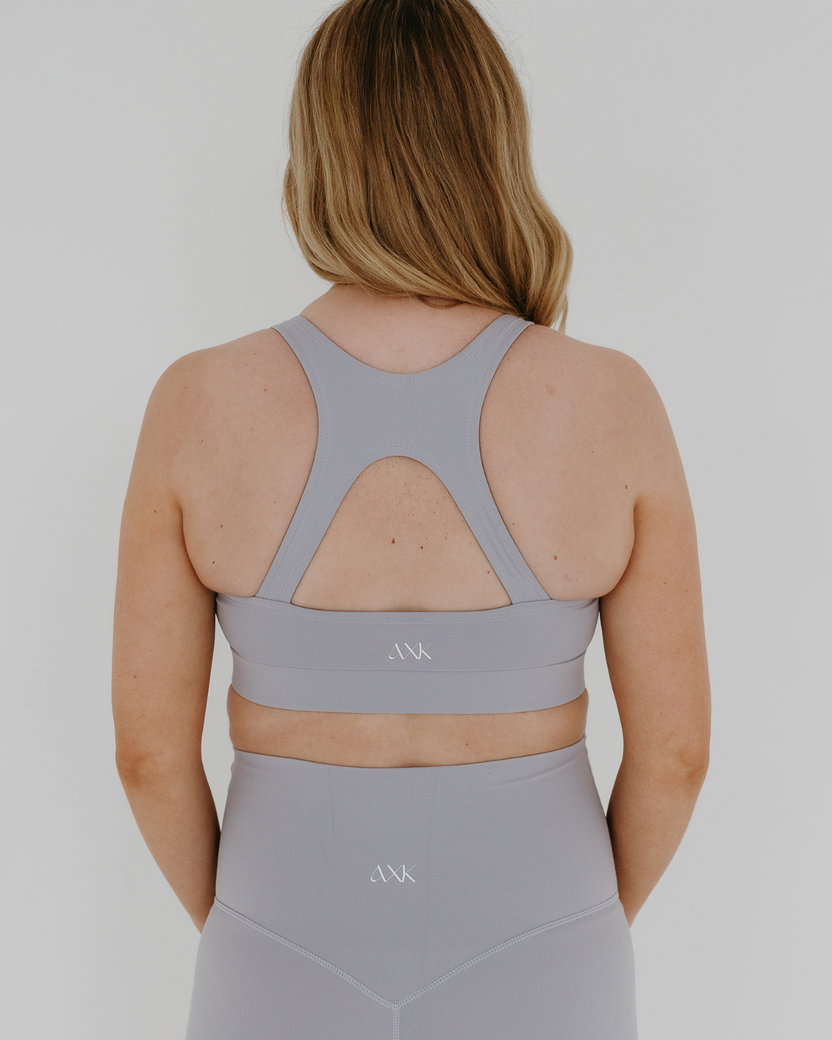 Back view of a woman wearing a light-gray, high-impact, racerback maternity and nursing bra with clip-down straps, standing against a white background. AXK premium maternity activewear