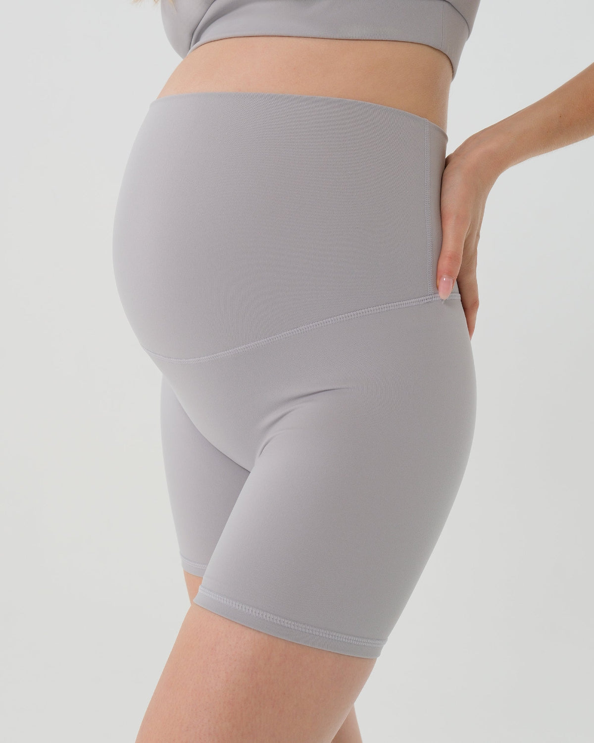 Pregnant woman wearing premium light grey maternity bike shorts with a supportive high waist and matching high-impact, racerback maternity and nursing sports bra, standing against a white background. Premium maternity athletic and activewear by AXK.