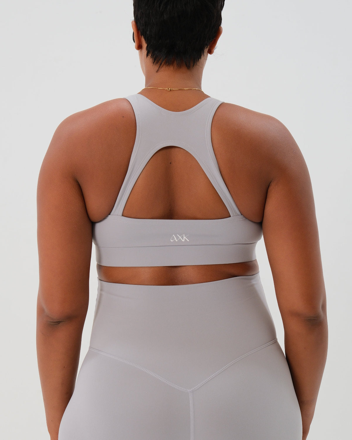 Back view of a woman wearing a light-gray, high-impact, racerback maternity and nursing bra with clip-down straps, standing against a white background. AXK premium maternity activewear