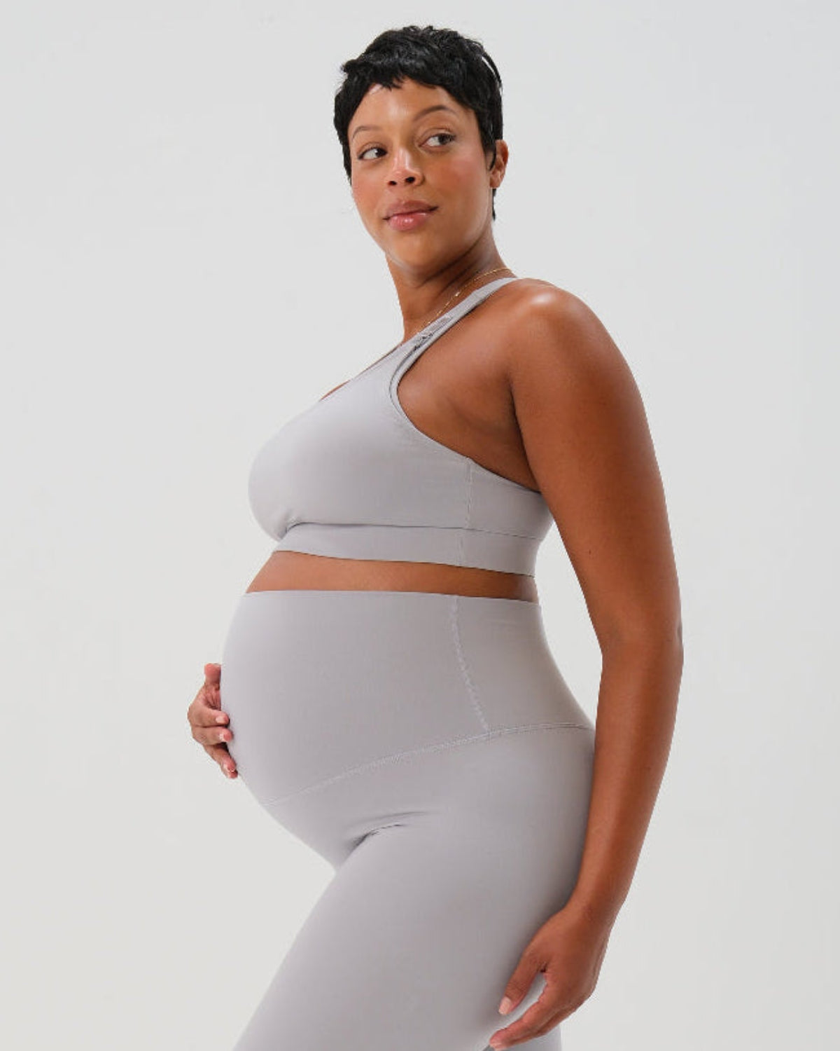 Side view of a pregnant woman wearing a light-gray, high-impact, racerback maternity and nursing bra with clip-down straps, standing against a white background. AXK premium maternity activewear