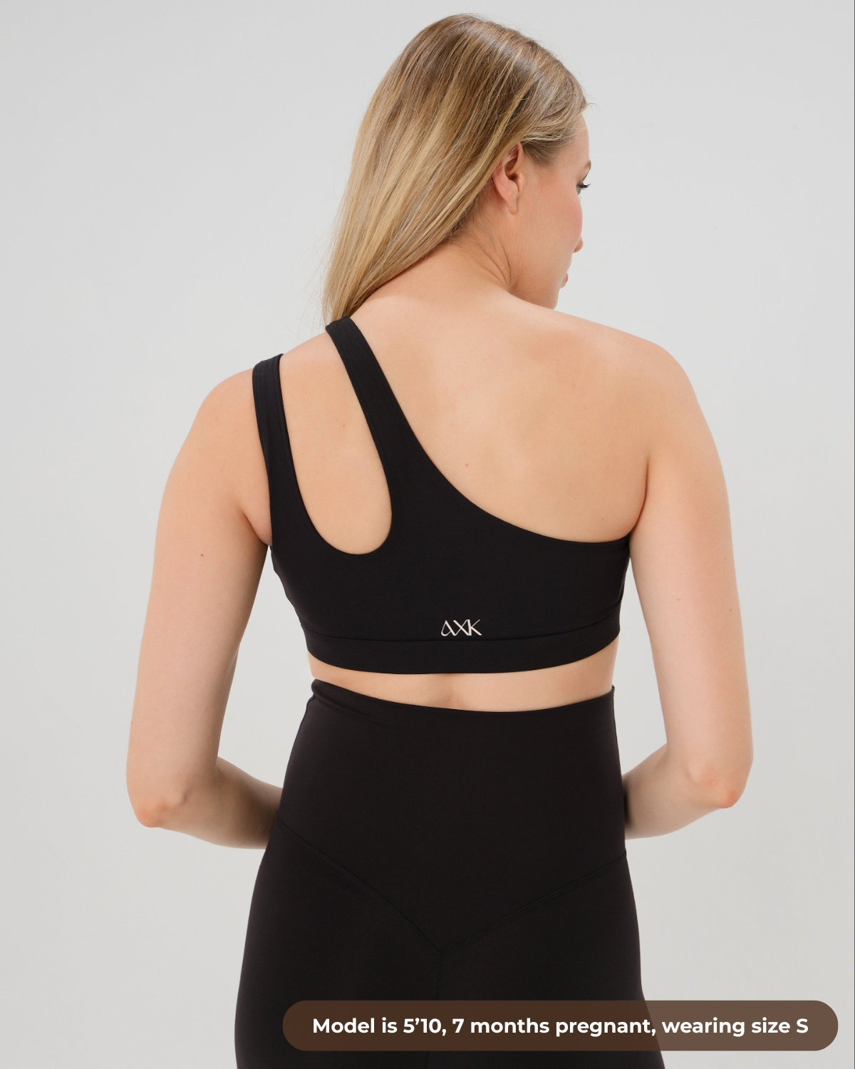 Pregnant woman in a one-shoulder black maternity and nursing sports bra, wearing matching black maternity leggings. Best selling 2026 premium maternity athletic and activewear by AXK, offered in regular and plus sizes, with glowing 5 star reviews. 